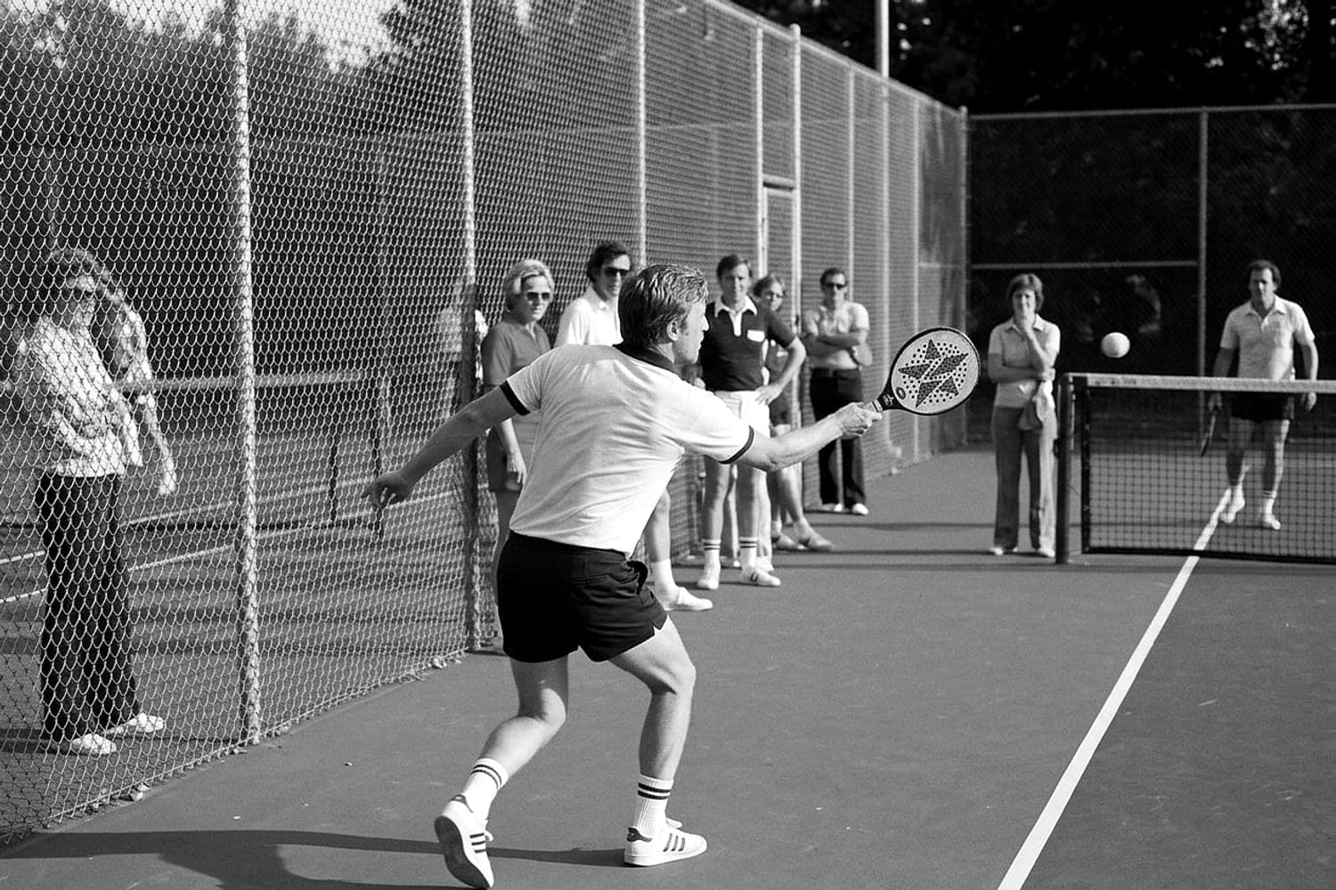 A man in tennis whites returns a shot on a platform tennis court while spectators stand along the sideline in daylight.